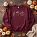 A burgundy crewneck sweatshirt with a white line-art graphic of a leaf, a hot cocoa cup, and a pumpkin, laid flat on a dark wood background surrounded by autumn leaves, pumpkins, a mug of whipped drink, and a knitted blanket.