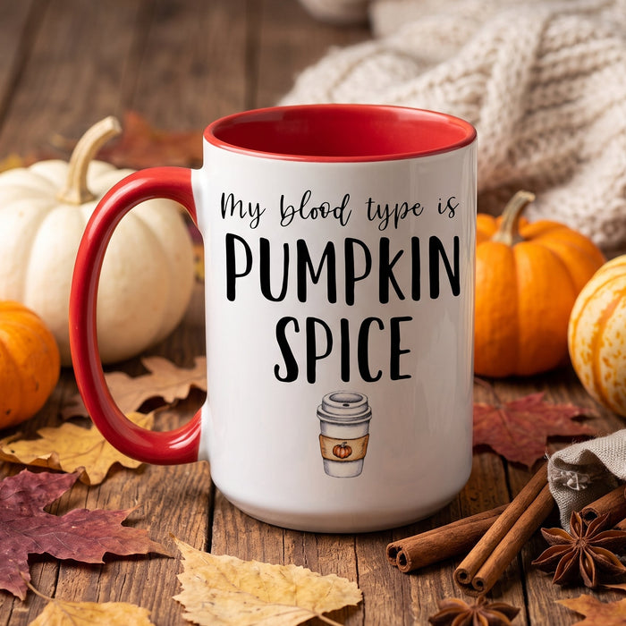 A white ceramic coffee mug with a red handle and red interior, printed with the phrase 'My blood type is PUMPKIN SPICE' and a small coffee cup graphic. The mug is styled on a rustic wooden surface with pumpkins, autumn leaves, cinnamon sticks, and a knitted blanket in the background.