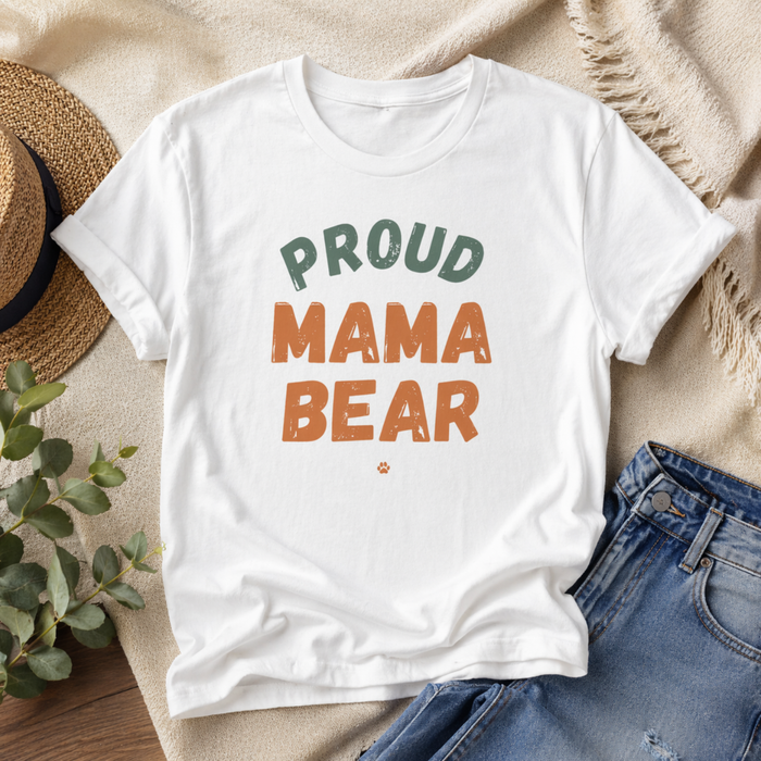 White t-shirt with 'Proud Mama Bear' text on a wooden surface with denim jeans and a hat.