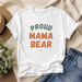 White t-shirt with 'Proud Mama Bear' text on a wooden surface with denim jeans and a hat.