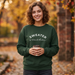 A woman stands outdoors on a cobblestone path in autumn, wearing a dark green crewneck sweatshirt with the words 'SWEATER weather' printed in white across the chest, and holding a coffee cup.